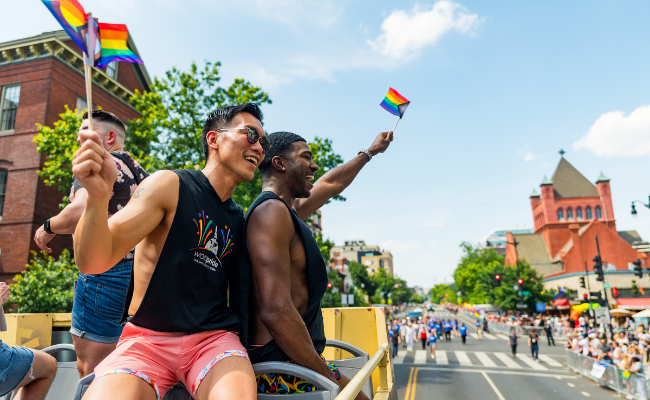 Two men in shorts and tank tops wave vibrant rainbow flags, celebrating pride and diversity in a lively atmosphere.