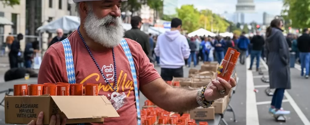 A man with a white beard and a blue checkered hat and suspenders, handing out orange beer cans at an Oktoberfest event in Washington D.C., with the U.S. Capitol building visible in the background.