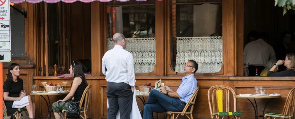 Diners relax with drinks and meals at an outdoor French café with lace-curtained windows and pink awning on a DC street.