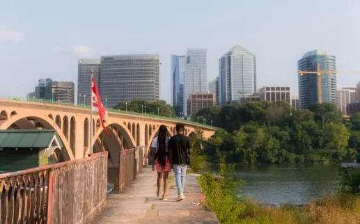 A couple walks along the Georgetown riverfront with a bridge and building of Arlington visible on the other side of the river. 
