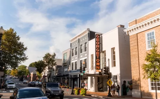 Pedestrians and cars pass by storefronts under a bright Georgetown sign on a bustling street in the historic Georgetown neighborhood of Washington, DC.
