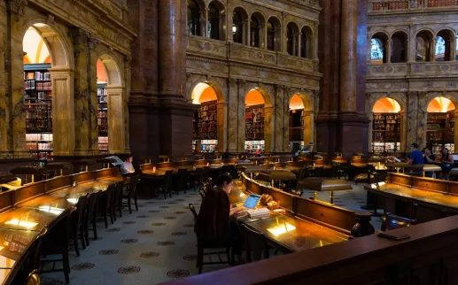 Visitors study and explore the grand marble reading room at the Library of Congress surrounded by arched alcoves and bookshelves.